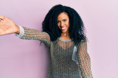 Orta yaşlı Afro-Amerikan kadın günlük giysiler içinde kameraya bakıp kucaklaşmak için gülümsüyor. Mutluluğu kucaklayan neşeli bir ifade.. 