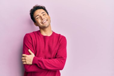 Yakışıklı İspanyol bir adam pembe arka plan üzerine günlük kıyafetler giyip kollarını kavuşturup kameraya bakarken gülümsüyor. pozitif kişi. 