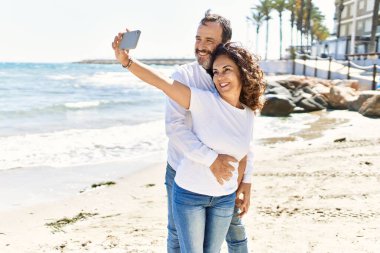 Orta yaşlı İspanyol çift mutlu gülümsüyor ve sahilde telefon başında selfie çekiyor..