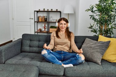 Orta yaşlı beyaz kadın yoga yapıyor ve kulaklık takıyor. Evdeki kanepede oturuyor..