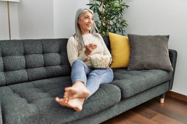 Orta yaşlı, kır saçlı, gülümseyen, kendine güvenen, evde film izleyen bir kadın.