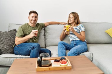 Genç İspanyol çift mutlu bir şekilde gülümsüyor. Evdeki kanepede oturup kahvaltı yapıyorlar..