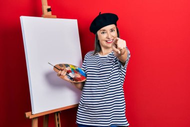 Orta yaşlı İspanyol kadın palet üstüne palet çiziyor. Resim sehpası seni ve kamerayı işaret ediyor. 