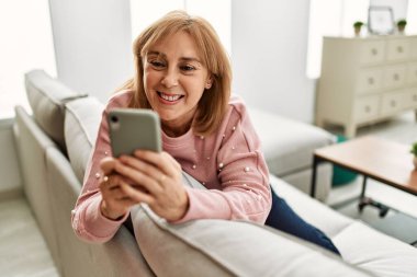 Orta yaşlı sarışın kadın akıllı telefon kullanıyor. Evdeki kanepede oturuyor..