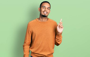 Genç, Afro-Amerikalı, günlük kıyafetler giyen, yüzünde kocaman bir gülümseme olan, elini kaldırıp kameraya bakan bir adam.. 