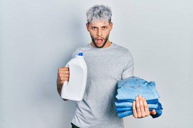 Çağdaş boyalı saçlı genç İspanyol bir adam çamaşırhane için kot pantolon ve şok olmuş yüzüne deterjan şişesi tutuyor. Şüpheci ve alaycı görünüyor. 