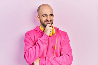 Spor kıyafetleri giyen ve eli çenesinde kulaklık takan genç kel bir adam soru sormayı düşünüyor, düşünceli bir ifade. Düşünceli bir yüzle gülümsüyor. şüphe kavramı. 