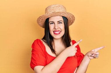 Genç İspanyol kadın yaz şapkası takıyor gülümsüyor ve kameraya bakıyor iki eliyle ve parmaklarıyla kenarı işaret ediyor. 
