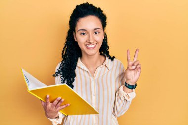 Kıvırcık saçlı, gülümseyen ve parmaklarını zafer işareti yaparken gösteren genç İspanyol kadın. İki numara.. 