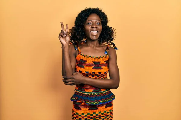 若いアフリカ系アメリカ人の女性が伝統的なアフリカの服を着て 勝利の兆候を行うカメラで顔をウィンク幸せな笑顔で — ストック写真