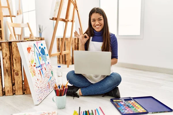 年轻的黑发女子坐在艺术工作室的地板上 手握笔记本电脑 手握手签 笑容可亲地摆出一副极好的手势 — 图库照片