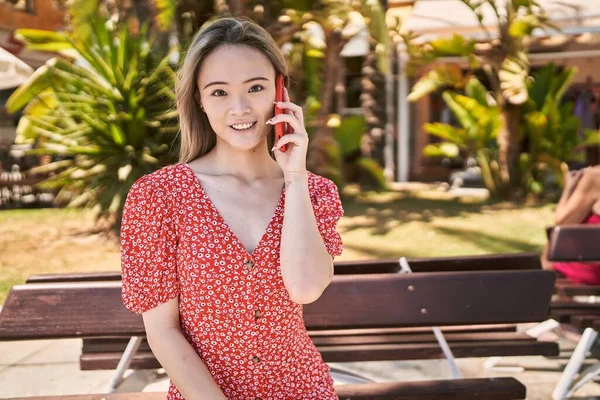 Genç Çinli kız gülümsüyor. Akıllı telefonda konuşuyor. Kentteki bankta oturuyor..