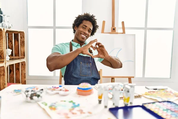 アートスタジオのテーブルに座っている若いアフリカ系アメリカ人男性は ハートのシンボルと手で形を見せるのが大好きです ロマンチックなコンセプト — ストック写真