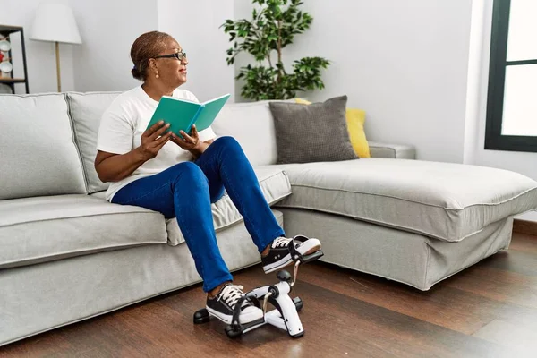 Senior african american woman reading book doing sport using bike pedals machine at home