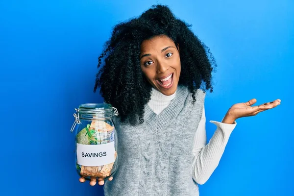 African american woman with afro hair holding savings jar with south african rands money celebrating achievement with happy smile and winner expression with raised hand