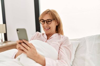 Orta yaşlı sarışın kadın evde yatağında akıllı telefon kullanıyor..