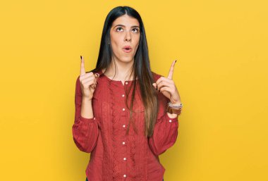 Gündelik kıyafetler giyen genç İspanyol kadın şaşırmış ve şaşırmış bir şekilde yukarı bakıyor ve parmaklarıyla işaret ediyor ve kollarını kaldırıyor.. 