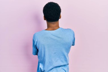 Genç, Afro-Amerikan bir adam, günlük mavi tişörtler giyiyor. Arkaya bakıyor, kollarını kavuşturuyor. 