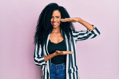 Orta yaşlı Afro-Amerikalı kadın, sıradan kıyafetler giyiyor. Elleriyle büyük ve büyük boy işaretleri gösteriyor. Ölçüm sembolü. Kameraya bakıp gülümsüyor. ölçüm kavramı. 