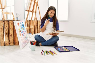 Sanat stüdyosundaki not defterine şüphe ifadesi çizen genç Latin kadın.