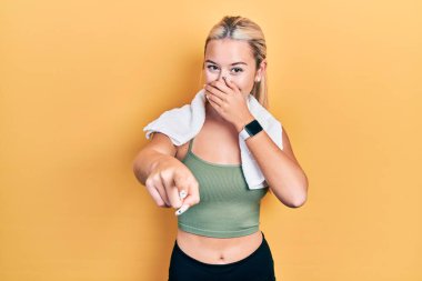 Spor kıyafetleri ve havlu giymiş genç sarışın kız sana gülüyor, eli ağzında kamerayı işaret ediyor, utanç ifadesi var. 