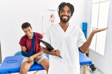 Kırık kollu bir adamla birlikte ağrı kesici kliniğinde çalışan genç İspanyol bir adam neşeli bir şekilde sunum yapıyor ve avuç içi kameraya bakıyor.. 