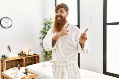 Kızıl saçlı, uzun sakallı bir adam sağlık merkezinde bornoz giyiyor gülümsüyor ve kameraya bakıyor iki eliyle ve parmaklarıyla yandan işaret ediyor.. 