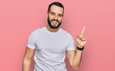 Sakallı, sıradan beyaz tişörtlü, yüzünde kocaman bir gülümseme olan genç bir adam. Elinle kameraya bakıyor.. 