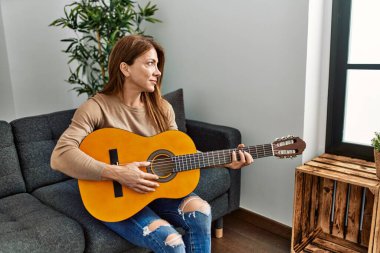 Orta yaşlı beyaz kadın klasik gitar çalıyor. Evdeki kanepede oturuyor..