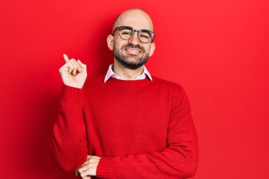 Genç kel adam, günlük kıyafetler ve gözlük takıyor. Yüzünde kocaman bir gülümseme var. Kameraya el ve parmak şeklinde bakıyor.. 