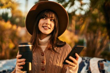 Kışlık şapkalı esmer kadın akıllı telefon kullanıyor ve parkta kahve içiyor.