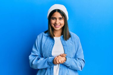 Tatlı yün şapkalı, elleri çapraz parmaklı, gevşemiş ve neşeli gülümseyen genç Latin kadın. Başarı ve iyimserlik 