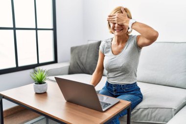 Orta yaşlı sarışın kadın evde dizüstü bilgisayar kullanıyor. Gülümsüyor ve elleri yüzünde gülüyor. Şaşırtmak için gözleri örtüyor. kör kavram. 