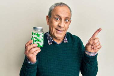 Yakışıklı, gri saçlı, elinde haplar olan mutlu bir şekilde gülümseyen, eli ve parmağı yana dönük yaşlı bir adam. 