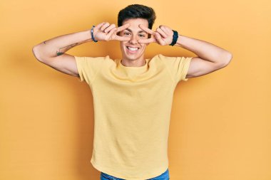 Gündelik sarı tişört giyen genç İspanyol adam barış sembolü yapıyor. Yüzünde parmaklar, gülümseyerek zaferi gösteriyor. 