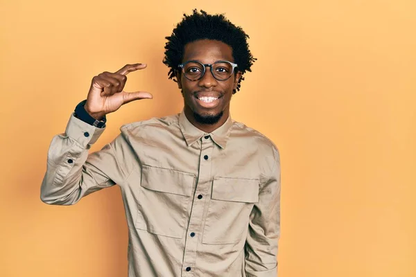 カジュアルな服と眼鏡を身に着けている若いアフリカ系アメリカ人の男性は 指を見てカメラと小さなサイズの看板を行う手で笑顔と自信を持ってジェスチャー 概念を測定し — ストック写真