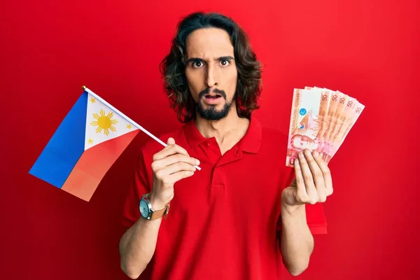 Young Hispanic Man Holding Philippines Flag Pesos Banknotes Shock Face — Stock Photo, Image