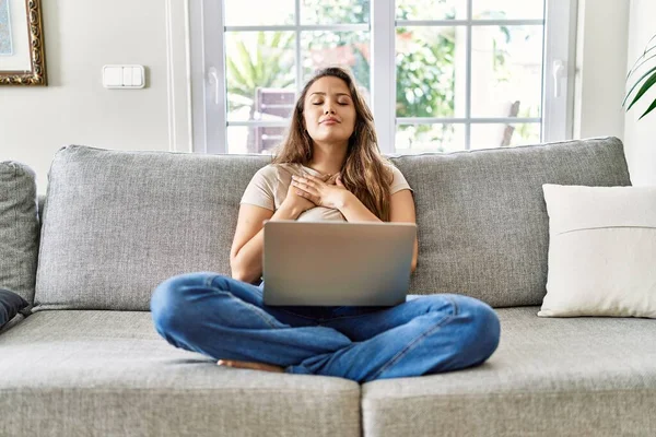 漂亮的年轻黑发女子坐在沙发上 在家里用电脑笔记本电脑微笑着 双手放在胸前 闭着眼睛 脸上挂着感激的手势 健康概念 — 图库照片