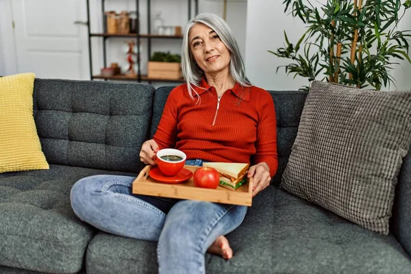 中年灰发女子坐在沙发上吃早餐 — 图库照片