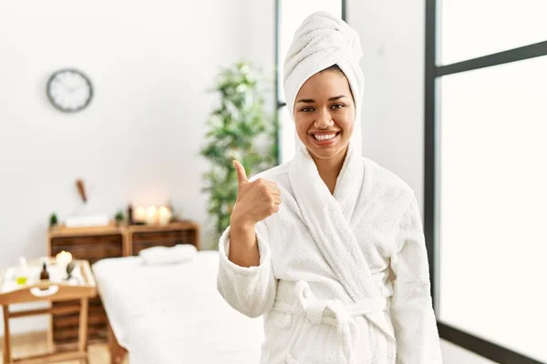 年轻的黑发女人穿着毛巾和浴衣站在美容中心 用手拿着快乐的大拇指做手势 赞成的表情看着相机显示的成功 — 图库照片