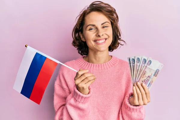 年轻的黑发女子举着俄罗斯国旗和卢布钞票 脸上挂着快乐而沉着的微笑 显示牙齿 — 图库照片