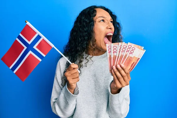 ノルウェーの国旗とノルウェーの紙幣を持っている若いラテン語の女性は怒っていると怒りで叫んで フラストレーションと激怒叫んで怒っている 怒りと攻撃的な考え方 — ストック写真
