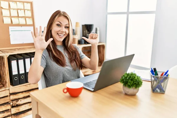 在办公室工作的年轻的黑发女人 拿着笔记本电脑 用手指指着六号 带着自信和快乐的笑容 — 图库照片