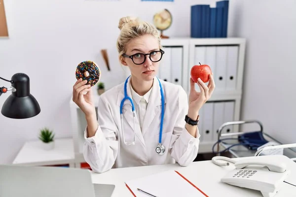 年轻的女医生拿着红苹果和甜甜圈在诊所里疑神疑鬼 神经质 皱着眉头因为问题而心烦意乱 消极的人 — 图库照片