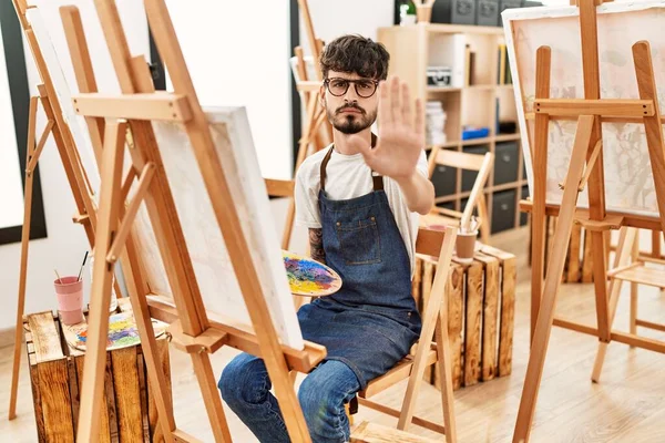Hispanic man with beard at art studio with open hand doing stop sign with serious and confident expression, defense gesture
