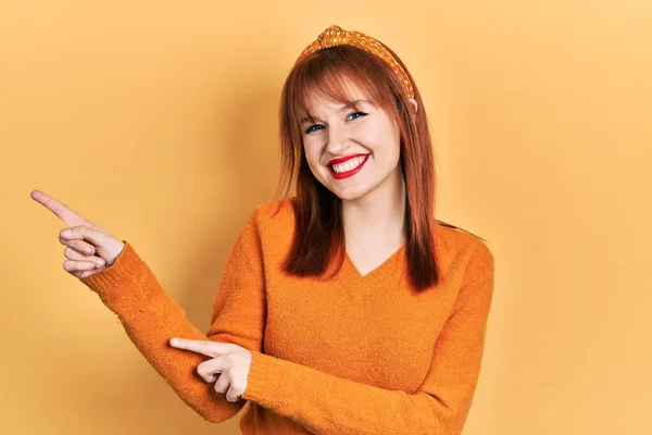 红头发的年轻女人穿着休闲的橙色毛衣 微笑着看着摄像机 用双手和手指指向旁边 — 图库照片