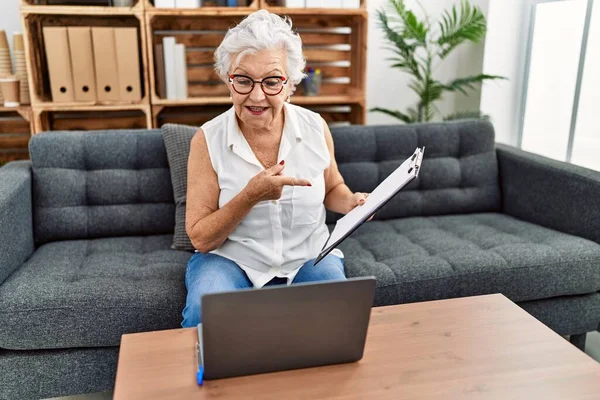 在咨询处工作的白发苍苍的老年妇女一边做在线治疗一边高兴地指指点点着自己的手指头 — 图库照片