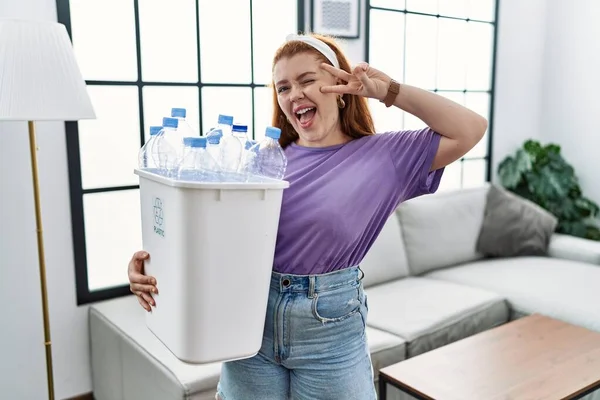Genç kızıl saçlı kadın elinde plastik şişelerle çöpleri geri dönüşüm sepetini tutuyor. Barış sembolü yapıyor. Yüzünde parmakları var. 