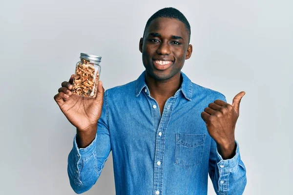 Joven Hombre Afroamericano Sosteniendo Frasco Con Nueces Apuntando Pulgar Hacia — Foto de Stock