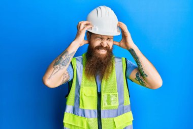 Uzun sakallı, güvenlik kaskı takan ve baş ağrısı çeken, çaresiz ve stresli bir ceketi olan kızıl saçlı adam. Ağrı ve migren yüzünden. Eller başının üstünde.. 